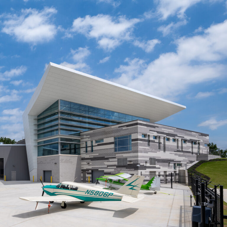 Exterior photography of the Chattahoochee Technical College Aviation Training Academy. The modern facility features a striking white cantilevered roof, expansive glass curtain walls, and multi-toned grey masonry. Two light aircraft are parked on the bright concrete tarmac in the foreground under a clear blue sky with scattered clouds.