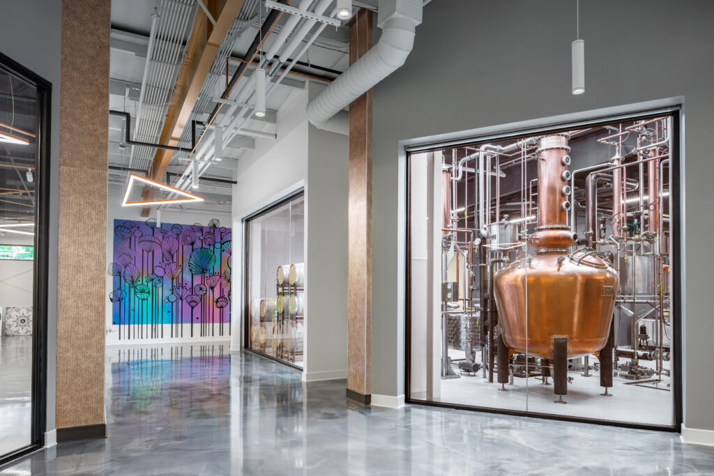 Architectural interior photography of the Distillery of Modern Art in Chamblee, GA. View through a large glass window into a production room featuring a large copper pot still and stainless steel piping. The foreground shows a polished concrete floor with reflections, gray walls, and a vibrant purple and blue modern art mural in the distance.