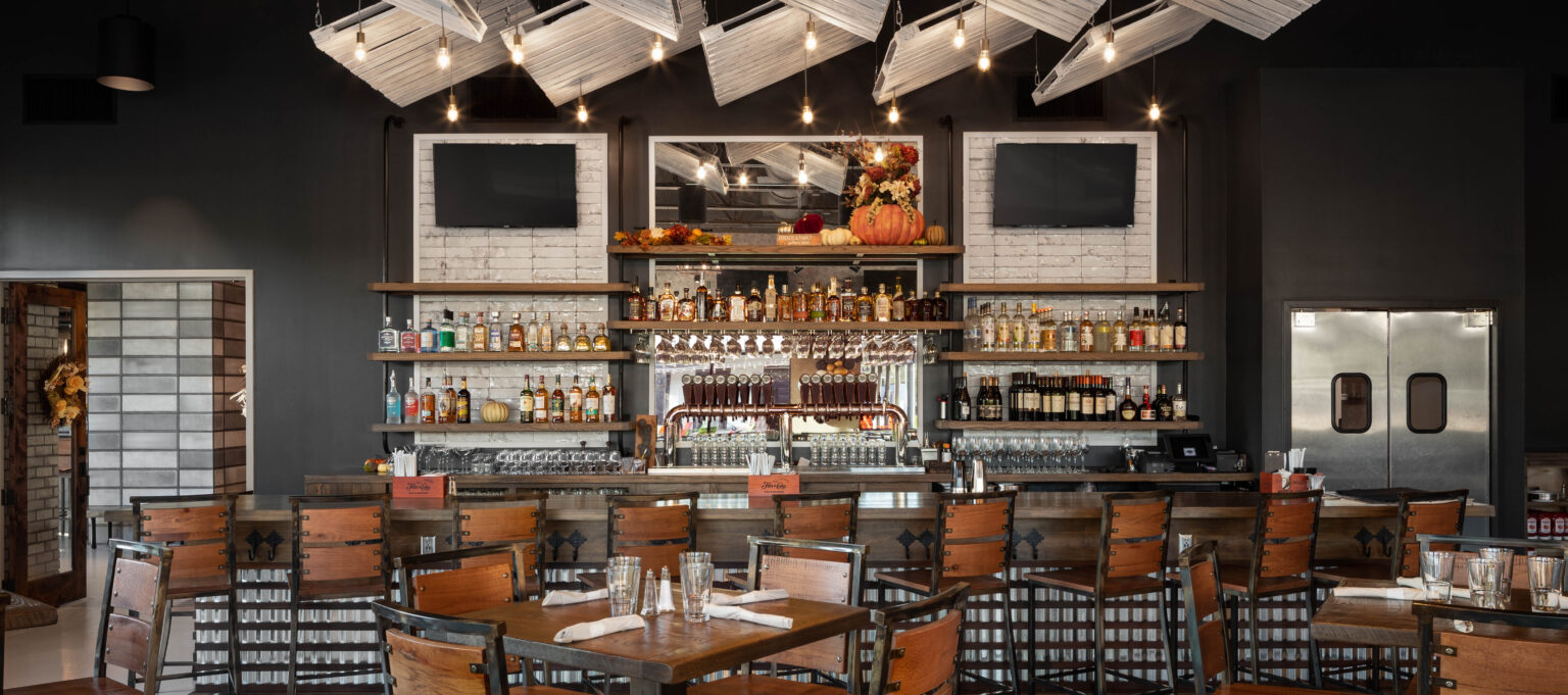 Architectural interior photography of Cherry Street Brewing at Halcyon in Alpharetta, GA. The shot features a symmetrical view of a modern industrial bar with a massive overhead lighting installation made of angled whitewashed wooden pallets and Edison bulbs. The foreground shows rustic wood and steel barstools, a dark gray wall, and a well-stocked back bar with white tile accents.